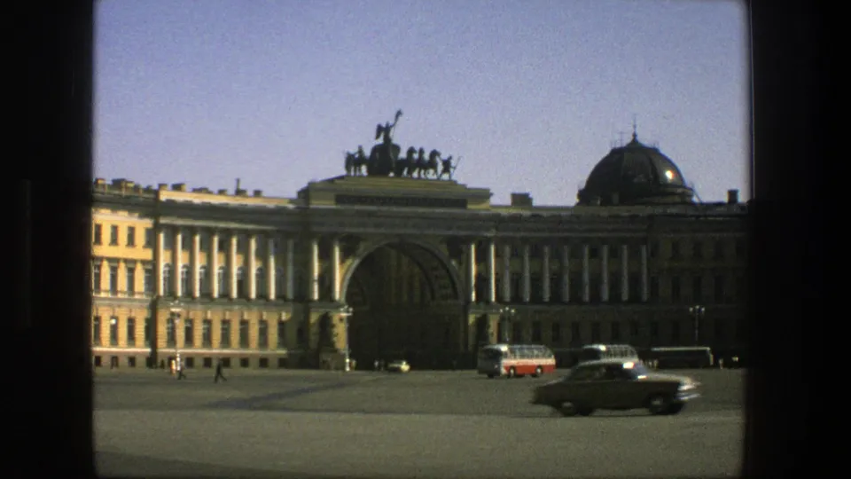 1975 Denmark street view with a dark car crossing the foreground, buses parked by a grand arched building, and pedestrians in the plaza.