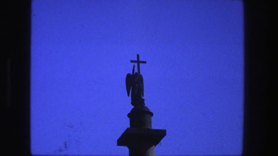 1975 Denmark 8mm frame showing the cross and winged figure lower in the frame, with blue sky and visible film texture.