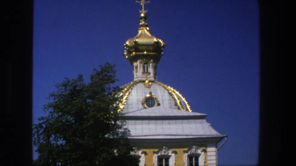 1975 Denmark 8mm frame with a gilded onion dome, cross, pale roof, yellow wall, and tree partially covering the church.