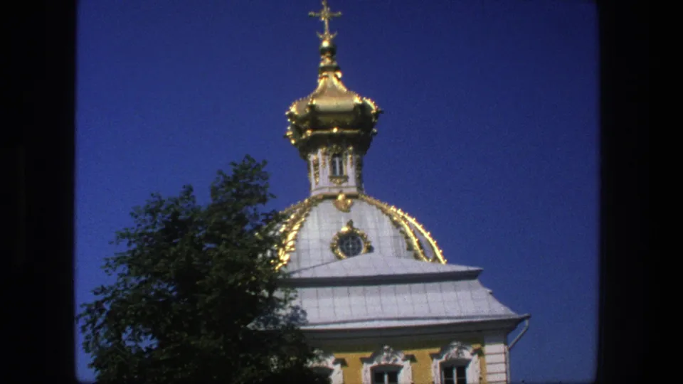 1975 Denmark 8mm frame of the ornate church dome and cross, partly obscured by tree foliage against a clear blue sky.