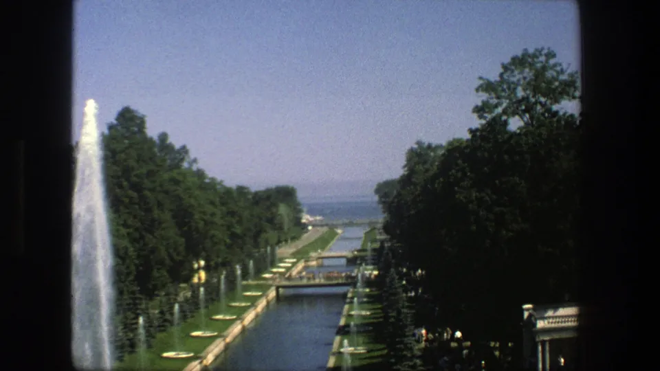 1975 Denmark 8mm view of a formal water channel with tall fountain spray, tree-lined paths, lawns, and distant water.