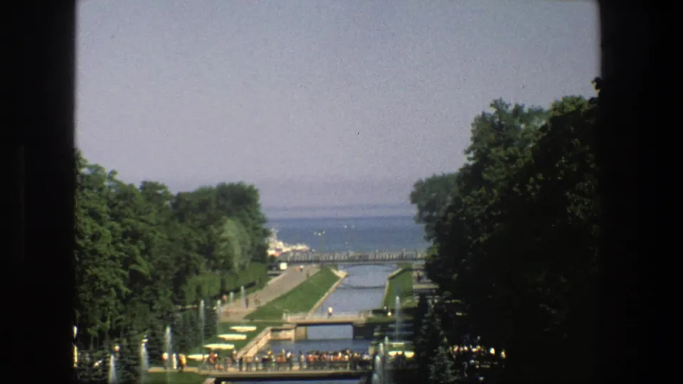 1975 Denmark home-movie view of a formal waterside garden where visitors gather near a bridge beside fountains and trees.