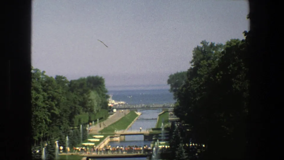1975 Denmark 8mm frame showing visitors beside a formal channel, low bridge, rows of fountains, trees, and distant open water.