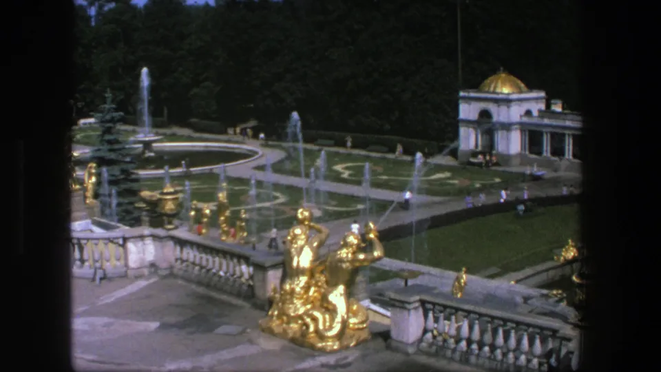 1975 8mm frame from Denmark showing a formal garden terrace with gold statuary, water fountains, clipped lawns, trees, and visitors.