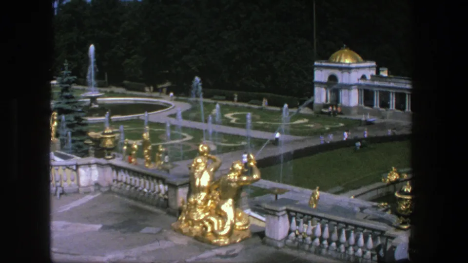 Wide 1975 Denmark garden view with gilded sculpture near a balustrade, active fountains, patterned lawns, a pavilion, and distant visitors.