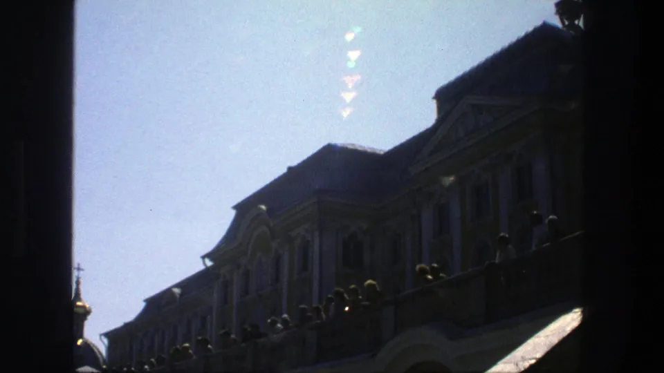 1975 Denmark home-movie frame of a grand facade, balcony viewers, dark doorway edges, and a pale sky after the light shift.