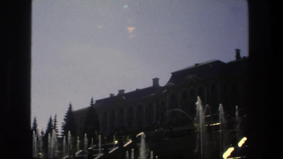 1975 Denmark 8mm frame showing a large ornate building behind active fountains, with evergreens and strong shadow across the lower frame.