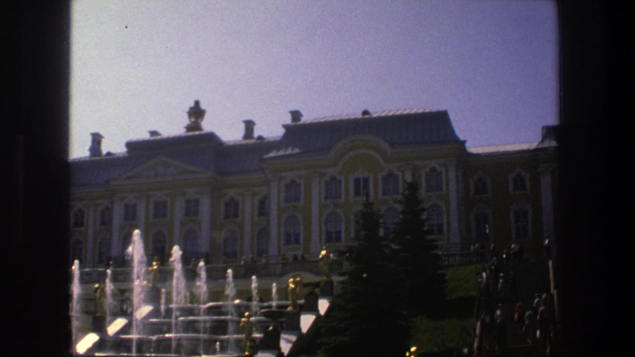 Vintage 8mm frame of the Danish palace grounds with fountain spray, lawn, and ornate facade under a clear sky.