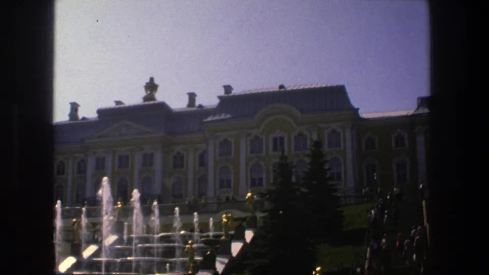Vintage 8mm frame of the Danish palace grounds with fountain spray, lawn, and ornate facade under a clear sky.