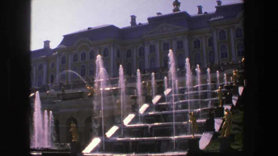 1975 8mm Denmark frame showing multi-tier palace fountains, gilded statues, a bright sky, and the ornate facade beyond.