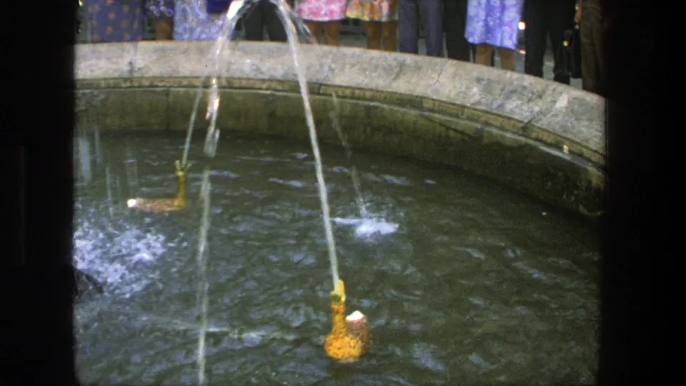 1975 Denmark home-movie frame showing golden mechanical ducks spraying water in a circular fountain with adult onlookers partly visible behind it.