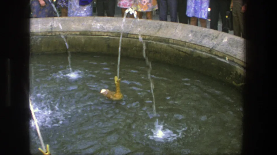 1975 Denmark home-movie still of a novelty duck fountain, with water splashing in the basin and adults visible behind the stone edge.