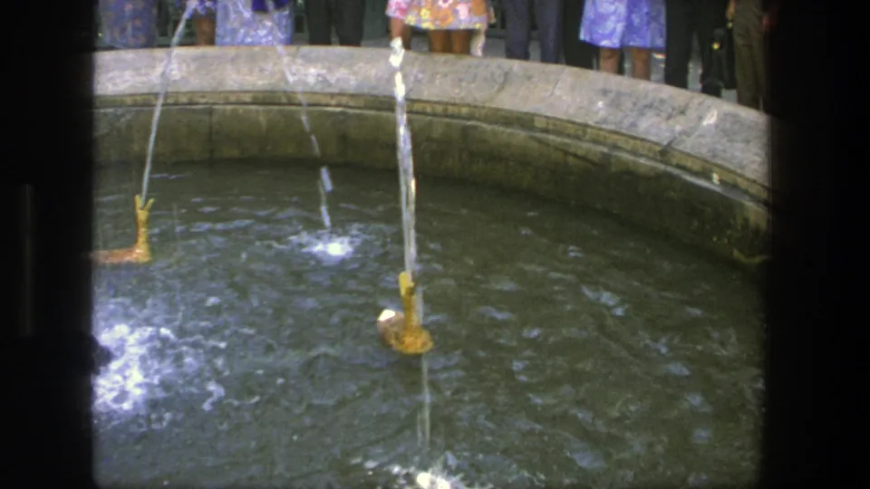 Late-frame 1975 Denmark view of a mechanical duck fountain, showing a vertical water spray, circular pool, and adult spectators in 1970s clothing.