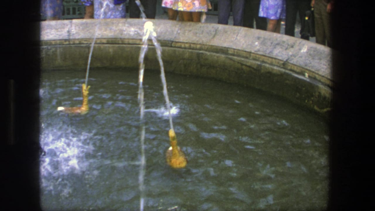 Round stone fountain basin with tan water spouts and visitors standing along the rim.