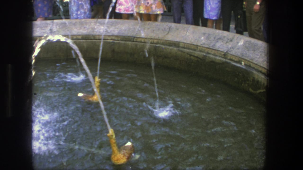 Tan fountain spout spraying across a round basin with people standing behind it.