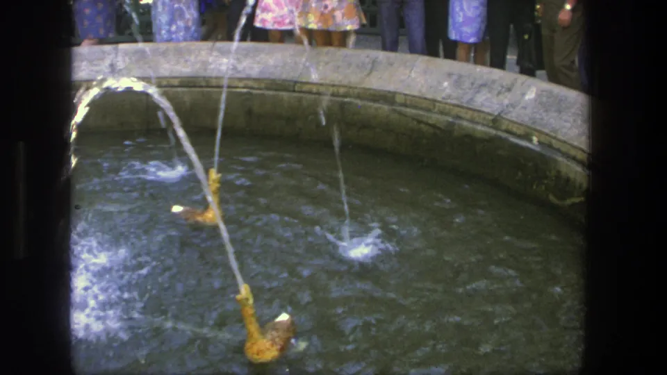 1975 Denmark 8mm frame of a tan fountain jet arcing over a circular basin, with visitors partly visible behind the rim.