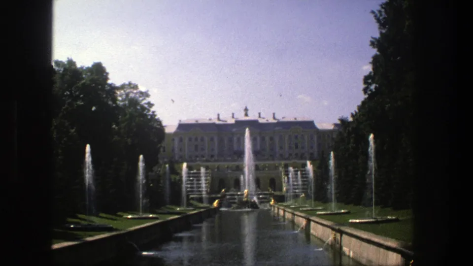 1975 Denmark 8mm frame of a long formal water garden with fountain jets, dark trees, lawns, and a palace-like building beyond.