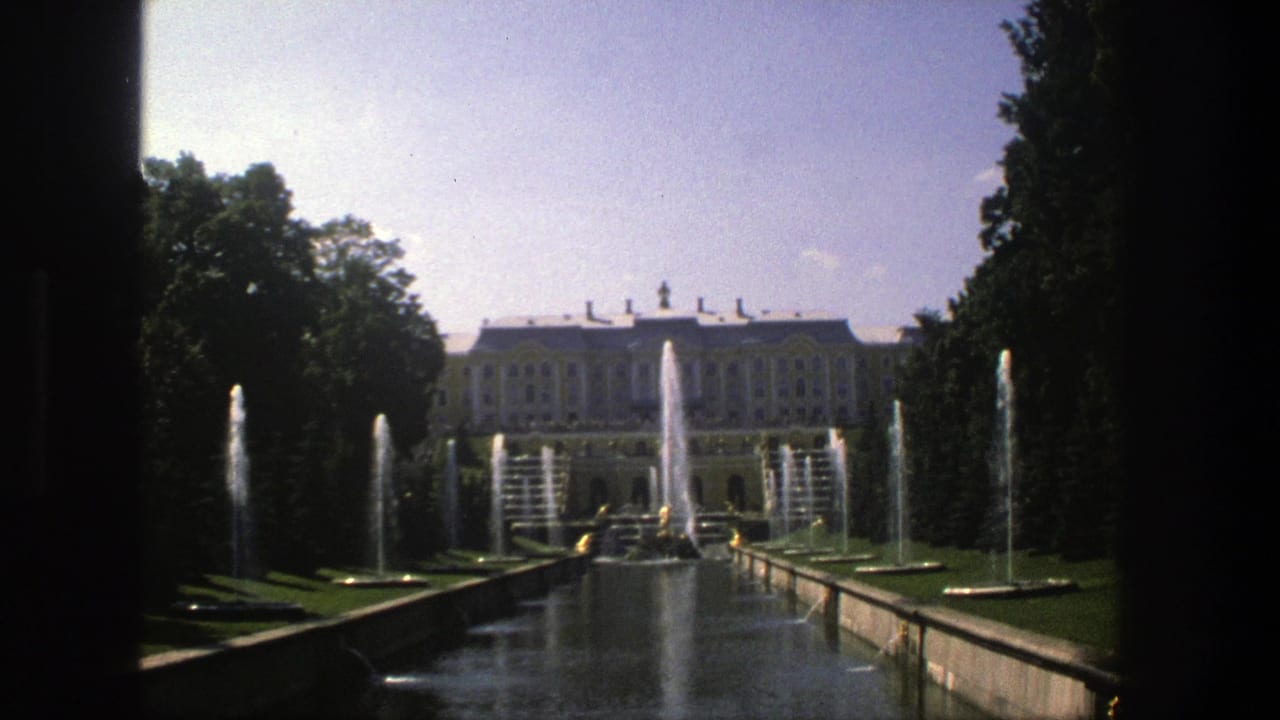 Wide 1975 Denmark travel frame of a palace garden axis with water jets, clipped lawns, trees, and a pale building beyond.