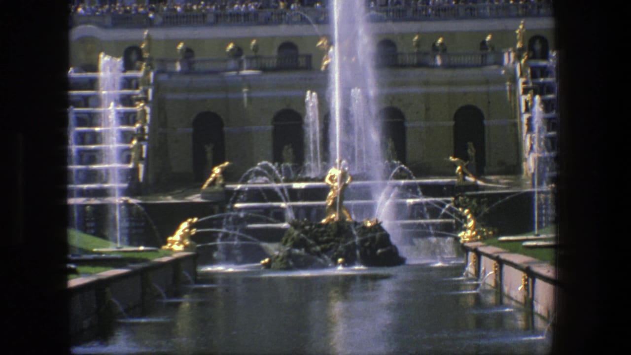1975 Denmark 8mm frame showing a grand fountain court with gilded male sculptures, tall water streams, a yellow arched facade, and visitors above.