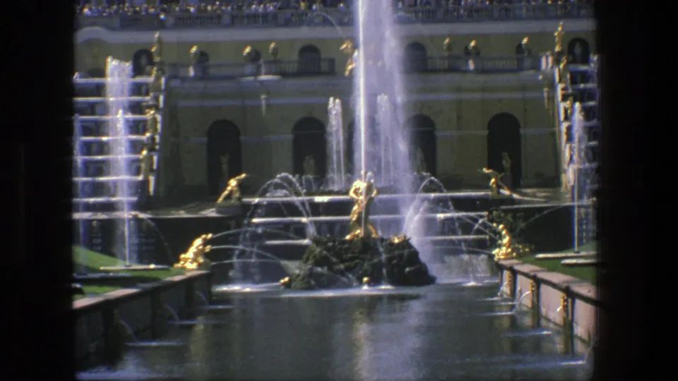 1975 Denmark 8mm frame showing a grand fountain court with gilded male sculptures, tall water streams, a yellow arched facade, and visitors above.