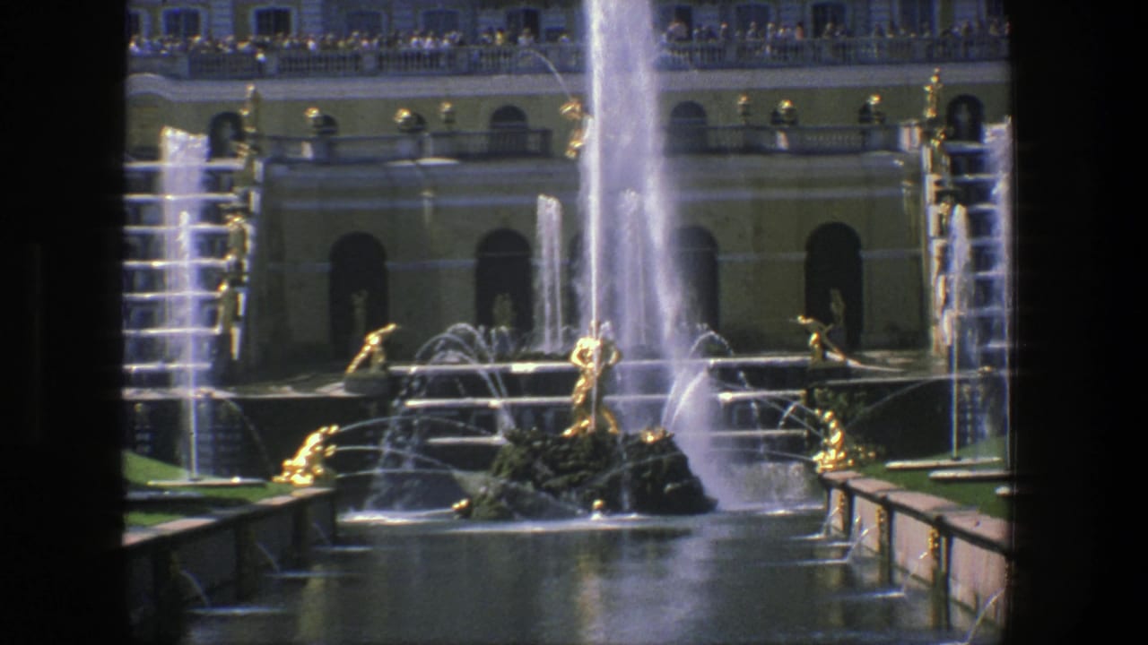 Fountain pool with cascading water, golden figures, classical facade, railings, and distant visitors on a balcony.