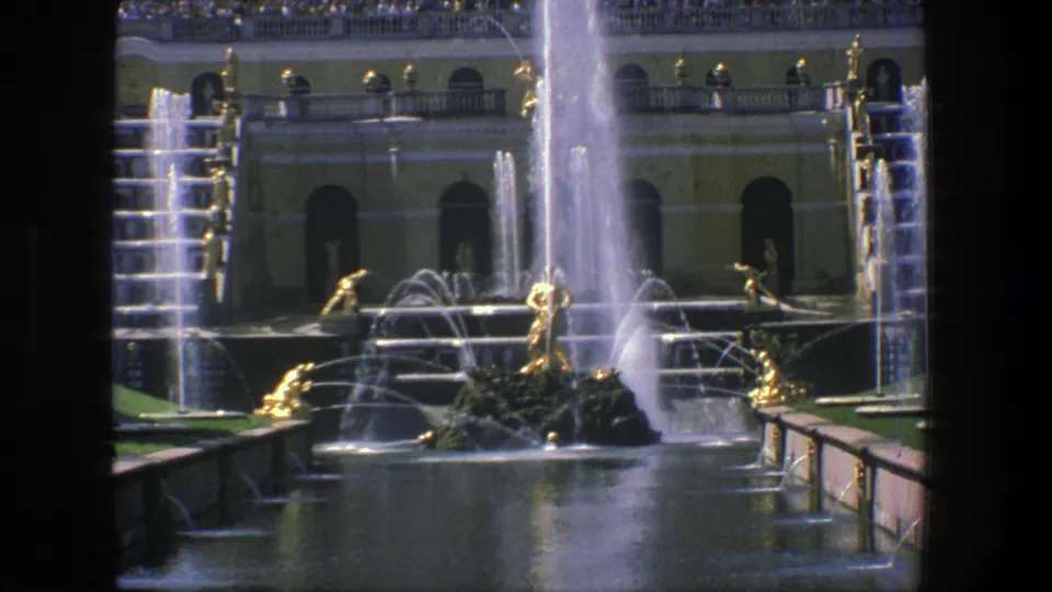 1975 Denmark 8mm still showing an ornamental fountain with arcing streams, gilded male sculptures, a yellow classical facade, and distant onlookers.