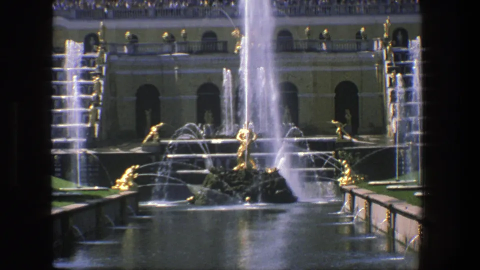 1975 Denmark home-movie frame with a grand fountain monument, gilded sculptural figures, bright water spray, pool reflections, and arched architecture.