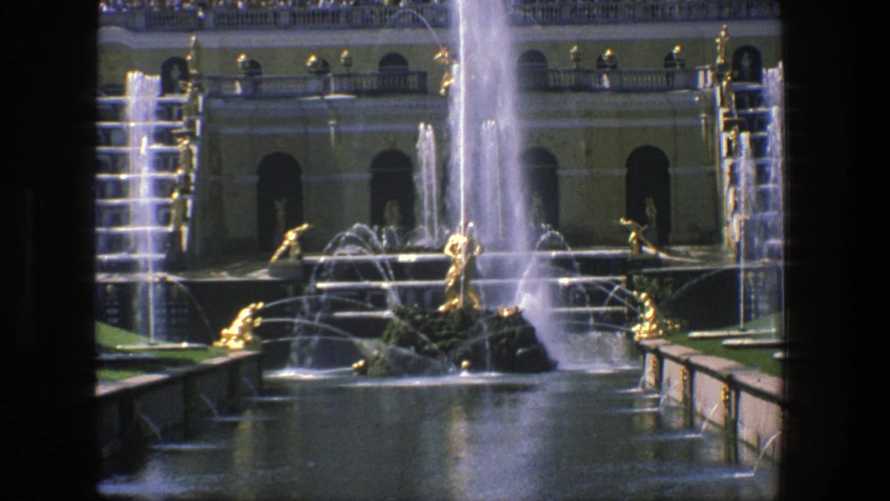 1975 Denmark-shot 8mm frame showing an ornate fountain with gold-toned statues, water jets, and a pale formal building behind it.