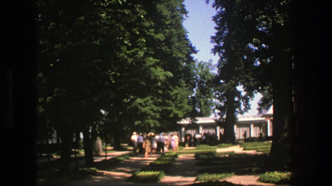 Tree-lined park path with visitors gathered near a light-colored pavilion.