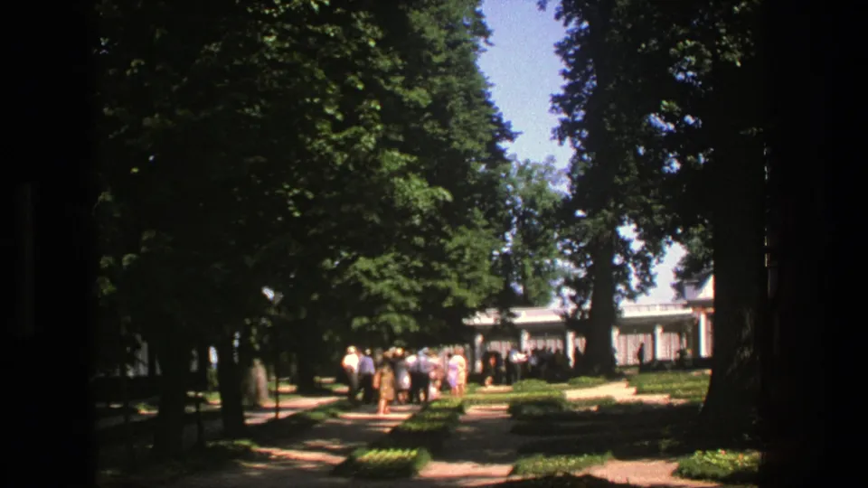 1975 Denmark-shot 8mm frame of visitors walking and gathering along a shaded garden path near a white pavilion-like structure.