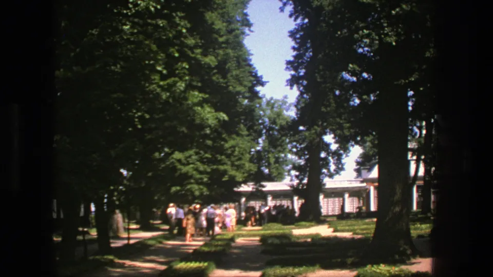 1975 Denmark-shot 8mm frame showing a group of visitors on a broad garden path beneath tall trees, with a light building in the background.