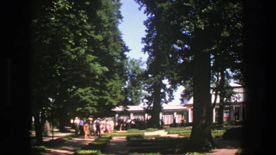 1975 Denmark-shot 8mm frame of visitors gathered on a tree-lined garden walkway with trimmed beds and a pale structure beyond.