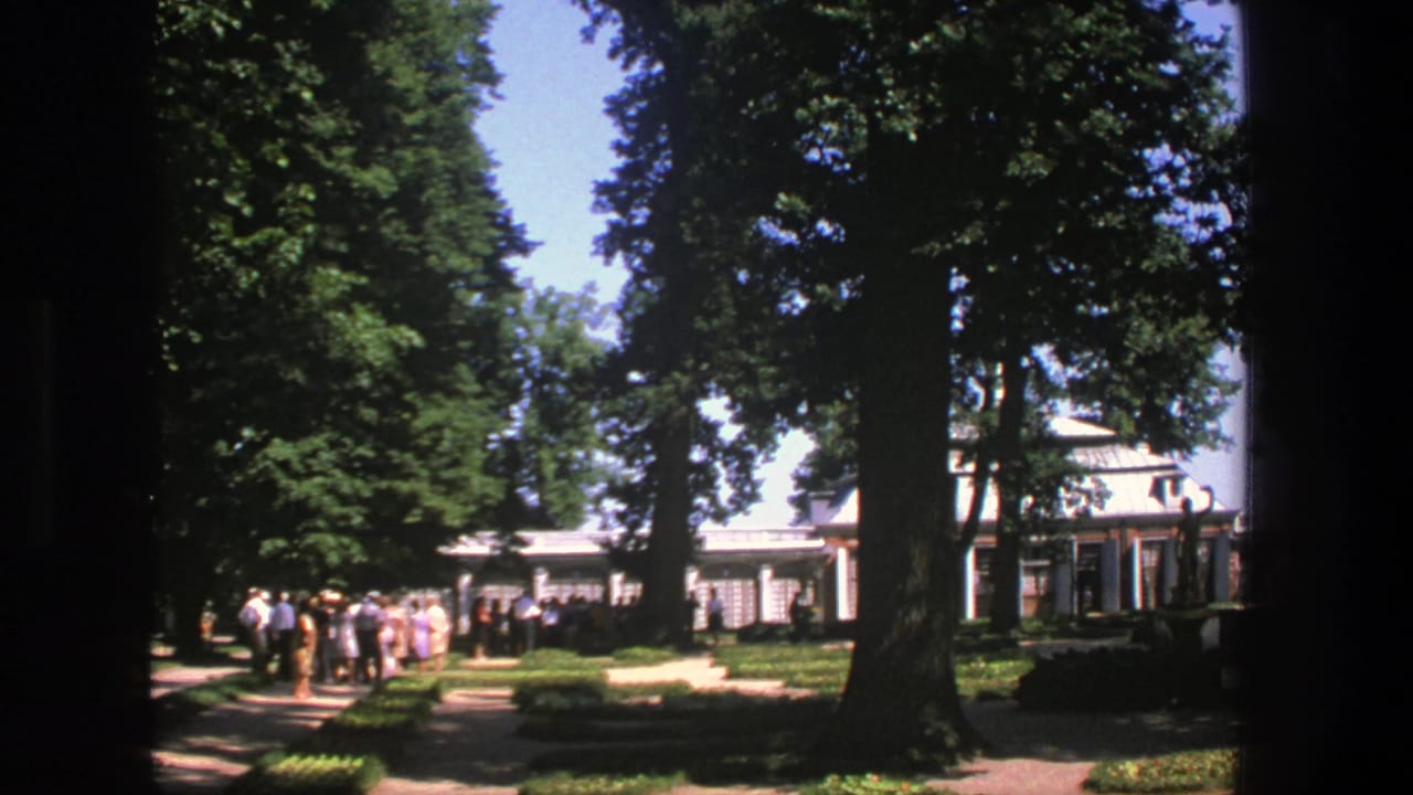 A group of people waits beside hedges and trees in front of a white building.