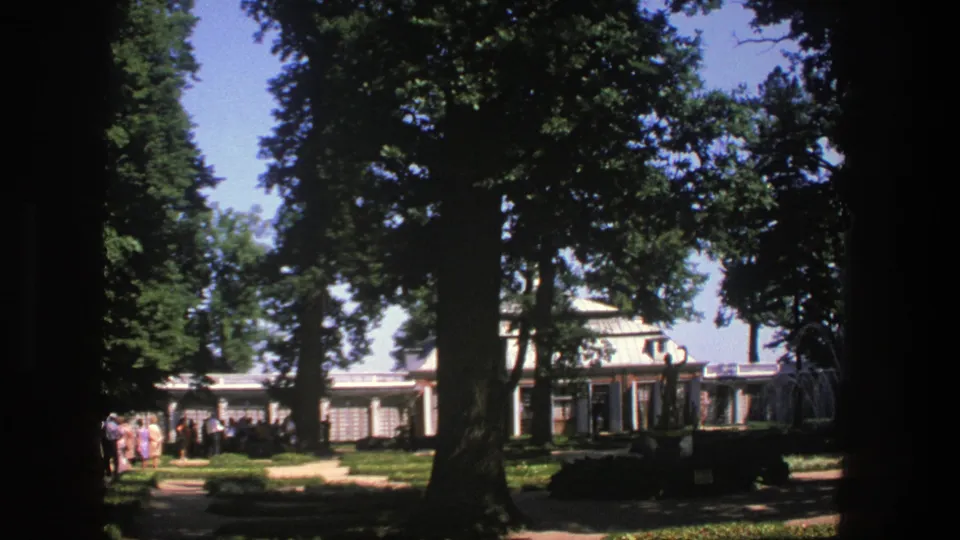 1975 Denmark home-movie frame with people in line at the left, a white building behind trees, and strong sunlight across the grounds.