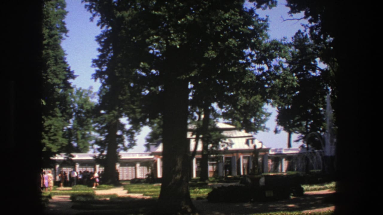 Shaded park view with tall trees, visitors, and a columned building in the background.