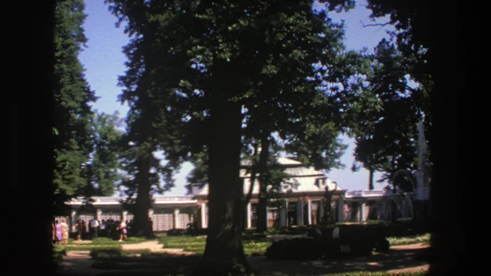 1975 Denmark home-movie frame showing tall trees, garden paths, visitors, and a classical-style building beyond the shade.