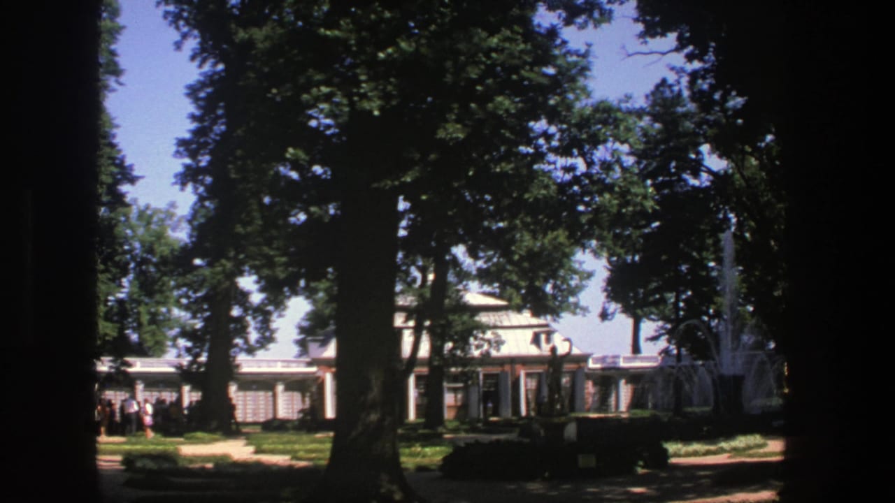 Trees and garden path frame a building with columns and a fountain spray at right.