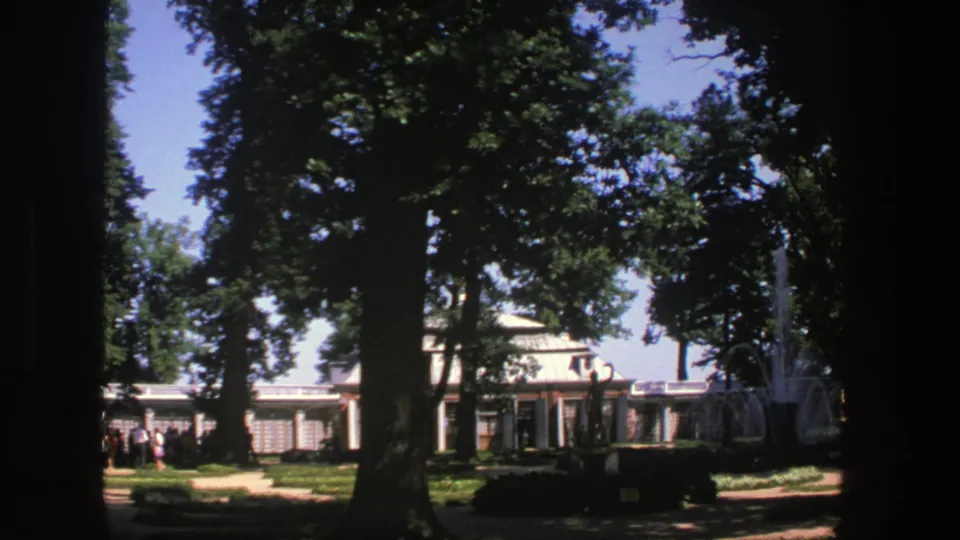 1975 Denmark frame with mature trees, a formal path, a columned building, distant visitors, and fountain spray on the right.