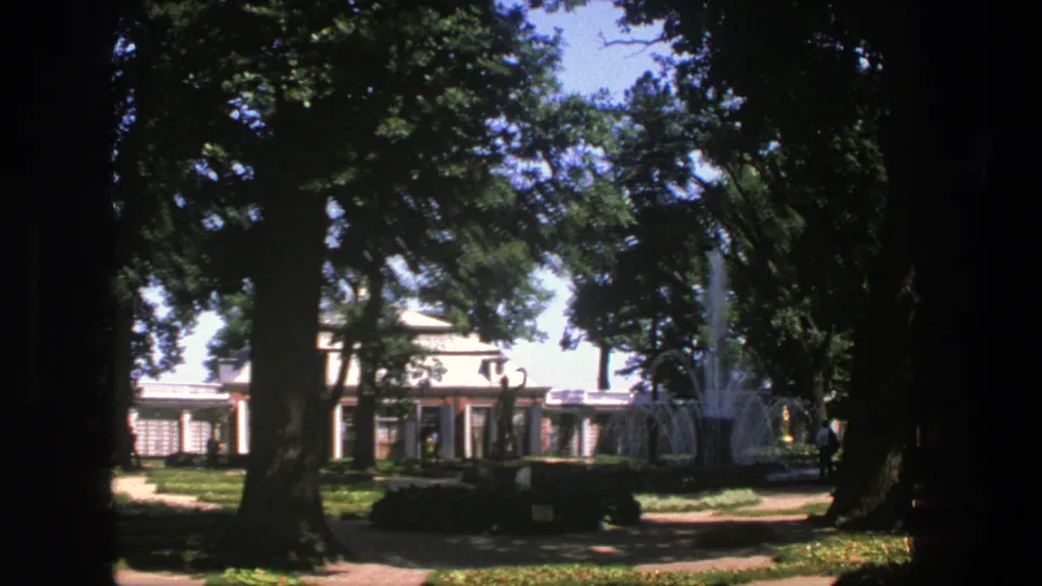 1975 Denmark frame showing a visitor-shot view across shaded trees toward a formal building and fountain in the grounds.