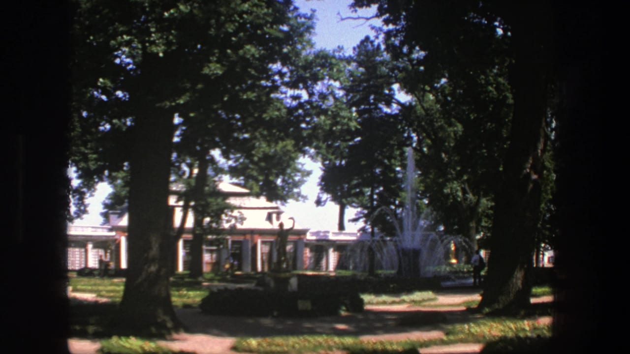 Shaded park path with large trees, a fountain, and a pale building in the background.