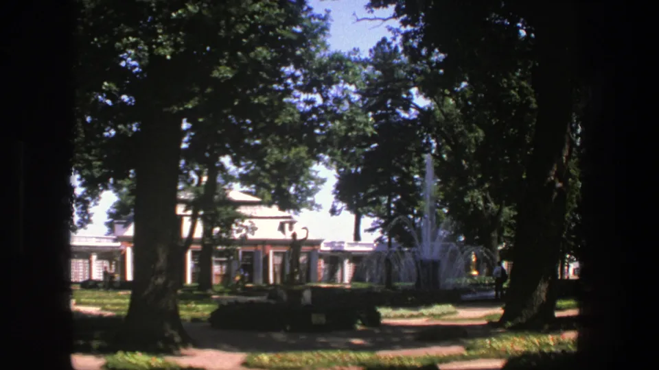 1975 Denmark home-movie frame showing a shaded park path, mature trees, a fountain, and a pale building beyond the greenery.