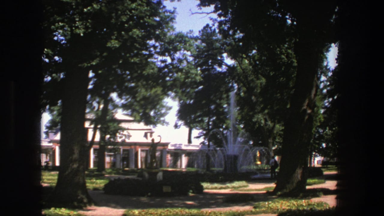 1975 Denmark park footage with tall trees, a fountain, a pale building, and a distant person near the path.
