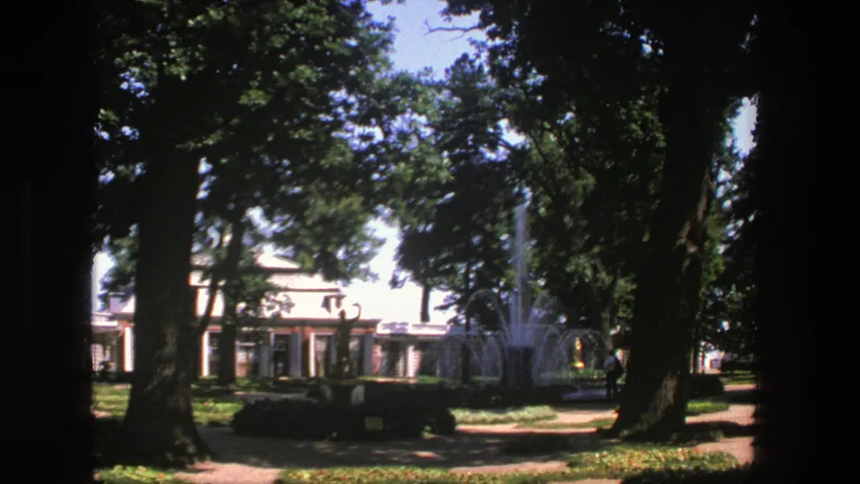 1975 Denmark park footage with tall trees, a fountain, a pale building, and a distant person near the path.