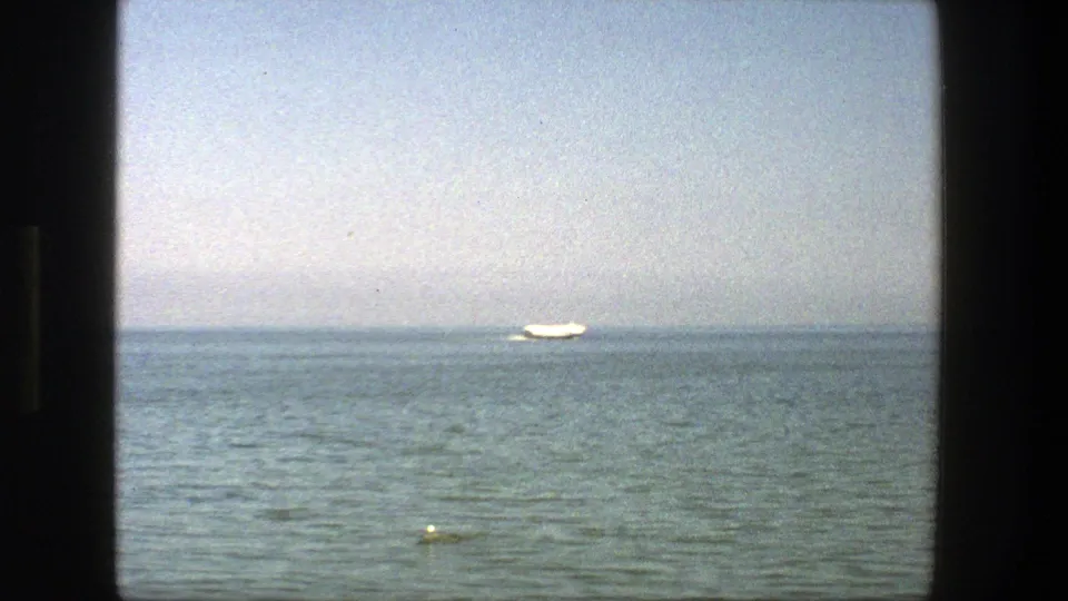 1975 Denmark coastal view on 8mm film, showing calm water, a distant white boat, horizon, and pale sky.