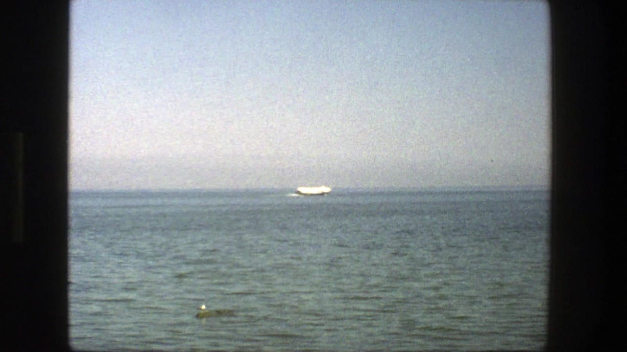 Distant white sailboat on calm water beneath a hazy sky, framed by dark film borders.