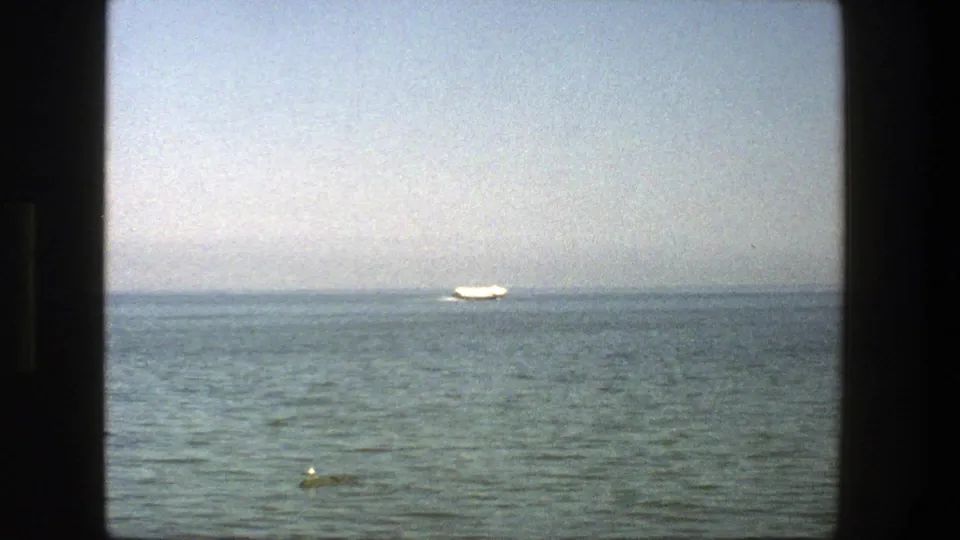 1975 Denmark 8mm frame showing a far white sailboat near the horizon with calm sea, hazy sky, and a small buoy in front.