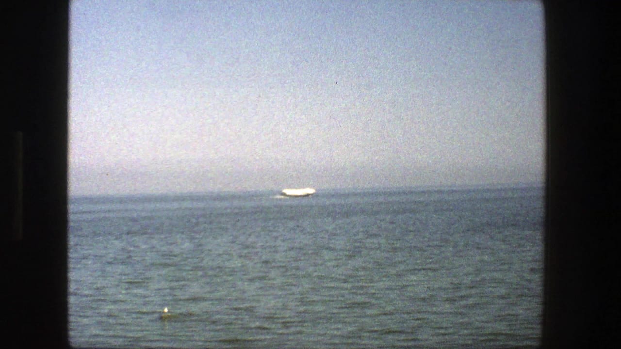 1975 Denmark home-movie frame with a distant white boat near the horizon, calm open water, pale haze, and dark border edges.