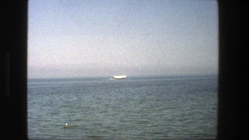 1975 Denmark home-movie frame with a distant white boat near the horizon, calm open water, pale haze, and dark border edges.
