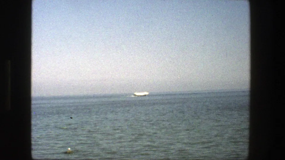 1975 Denmark 8mm view of a distant white sailboat on open water, with a buoy in the foreground and soft haze across the horizon.