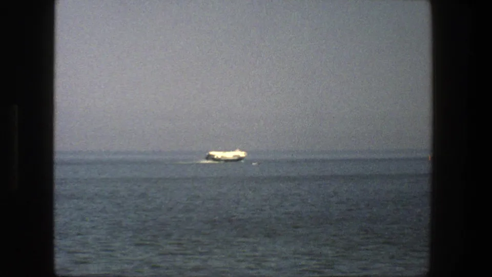 1975 Denmark home-movie frame showing a white vessel farther right on calm sea, with a faint wake and hazy horizon behind it.